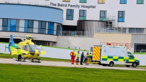 Yellow air ambulance and a person being taken into an ambulance, with a hospital in the background