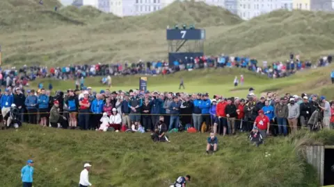 Reuters Crowds gather on a golf course behind a rope to watch the golf on the course. The golf course is surrounded by hills and there is a black metal structure with the number 7 on it.