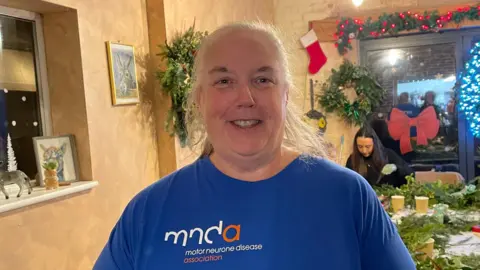 BBC/Amelia Riley Margaret Lidster - a woman with white hair tied in a ponytail and dressed in a blue T-shirt with the Motor Neurone Disease Association logo stands looking into the camera. Behind her, a group of people are sitting at tables making Christmas wreaths. Two completed wreaths and a garland also hang on the walls.
