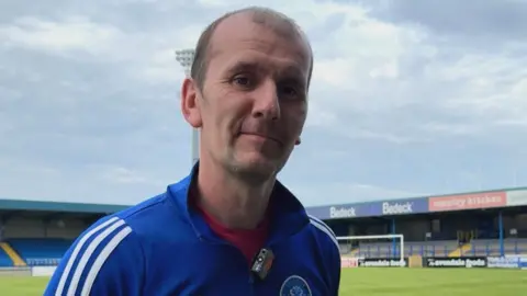 A man looks into the camera. He is wearing a blue tracksuit top with the Glenavon badge and white stripes on the sleeves, and a red t shirt. He has balding brown hair. A small football stand with blue seats and a football pitch is in the background.