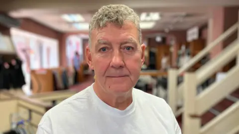 Sam Read/BBC Barry Day in a white T-shirt stands against the blurred background of the interior of Warwicks.