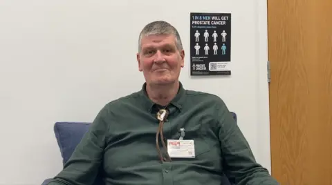 Humber and North Yorkshire Cancer Alliance Richard wears a green shirt and is sitting in a hospital ward besides a poster highlighting the main symptoms of prostate cancer.