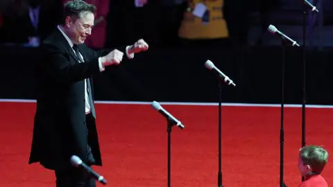 Getty photos Elon Musk with his son, X, on stage in Nasr Trump Rally in Washington, DC. Musk smiles and holds his hands. X face is invisible. 