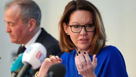 PA Media Emma Webber at a press conference. She is wearing glasses and is wearing a blue dress. She is sat in front of three press microphones.