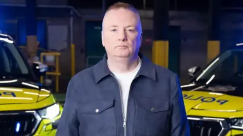 Stephen Nolan looking at the camera, he has brown hair. He is wearing a navy jacket that is zipped up halfway with a white t-shirt under it. Behind him are two police cars that have blue lights on.