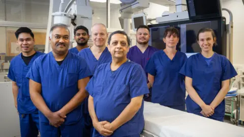 A group of six men and two women are all wearing blue scrubs and standing in an operating theatre