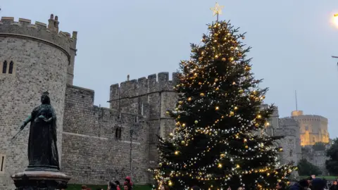 A highly-decorated Christmas tree can be seen in front of Windsor Castle under grey skies. The walls of the castle can be seen behind with some lit. A statue of Queen Victoria stands in front of the tree.