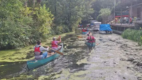 Steel Bones People in several turquoise canoes paddle along a river past moored boats and a quayside area, and trees on the opposite bank.