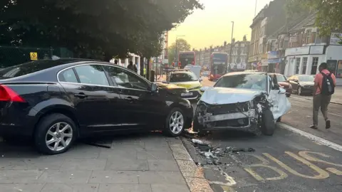 Two cars crashed into each other on a road and pavement with a police car and two buses behind them