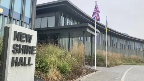 Brian Farmer/BBC The entrance to Cambridgeshire County Council's New Shire Hall headquarters. It is a long, sleek, grey building with tall windows and it looks modern. Grass has been allowed to grow tall in length out the front, which also has Union Jack and Ukraine flags on tall poles. There is a sign that says "New Shire Hall".
