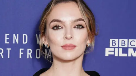 BBC News A headshot of Jodie Comer stood in front of a purple screen. She has has her hair tied up and i wearing small diamond hooped earrings and a black top and is looking into the camera.