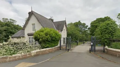The entrance to Jarrow Cemetery. A white two-storey house stands behind a metal fence on the left. The metal gate stands open providing access to the cemetery which is filled with trees.