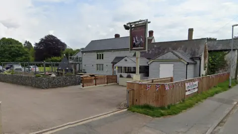 A car park is on the left of the picture with 4 cars. There is outdoor seating in front of the pub, with a wooden fence cornering it off. The pub is grey and has wooden cladding on the outside. It also has a sign outside that says British Queen on it. 