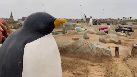 A mini golf course is covered in sand. Various features of the course are peeking out from under the sand including model boulders and large model penguins.