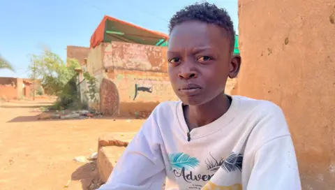 Ken Mugai / BBC Muzami, the 18 -year -old son of Intisar Adam Suleiman, looks directly at the camera lens with a sore expression. He wears a white sweat shirt and is sitting outside an orange building on a street in the Haj Yusuf district of the capital of Sudan Khartum - March 2025