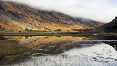 Gary Saddler Golden hills reflected in the water of a loch with a cloudy sky