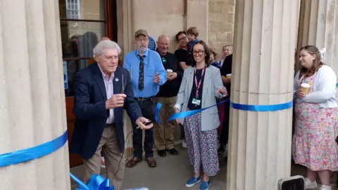 Paul Hayes/BBC A white-haired man in his 80s, wearing a dark blue jacket, a light blue shirt and light blue trousers, holds up a pair of scissors having just cut through a ceremonial blue ribbon tied between two columns at the ornate entranceway into a public building. Various other people stand around watching and applauding.