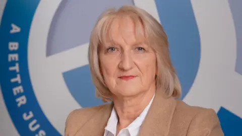 CJI Jackie Durkin. She has shoulder-length blonde hair with a fringe and blue eyes, wearing a white shirt and beige blazer.