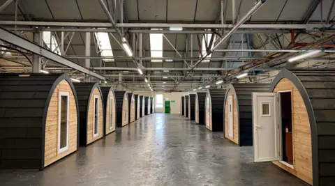 Tom Reece Two rows of wooden pods, like those found at campsites. Those pictured are inside a warehouse space. Each of the pods has a door, and the nearest on the right-hand side of the image is open.