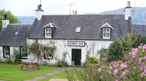 Colin Smith/Geograph The white-walled two-storey building has a garden in front of it with shrubs with pink flowers.