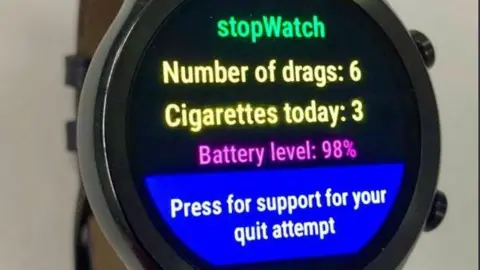 PA A smartwatch displaying the new app's messages. It shows the words stopWatch in green, with the message - Number of drags: 6, Cigarettes today: 3, Battery level: 98%, Press for support for your quit attempt. 