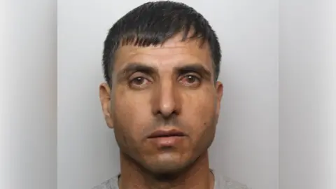 A police mugshot of a man with short black hair and wearing a grey top.
