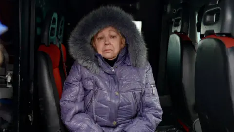 Goktay Koraltan/ BBC Olga sits inside a van wearing a thick warm purple coat with a fur hood, with a sad and worried expression on her face