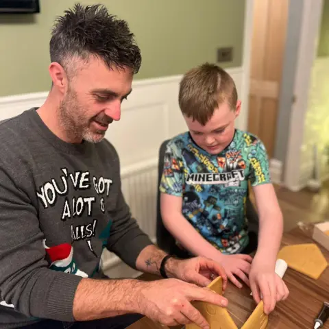 Nik Bannister A man with brown hair and a beard wearing a grey jumper with his son wearing a Minecraft top building a gingerbread house. 