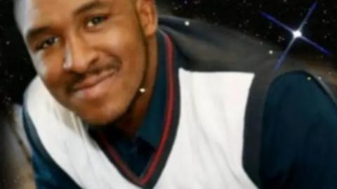 Dominic Wilson pictured in a white and black v-neck vest over a navy shirt, smiling. 