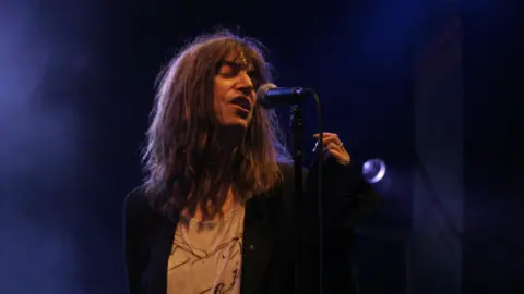 Leslie McGhie/WireImage A long-haired female musician onstage singing into a microphone. She is wearing a dark jacket and a light grey T-shirt