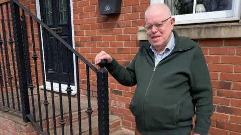 An elderly man in thinning white hair stands outside a red brick house holding on to a black railing