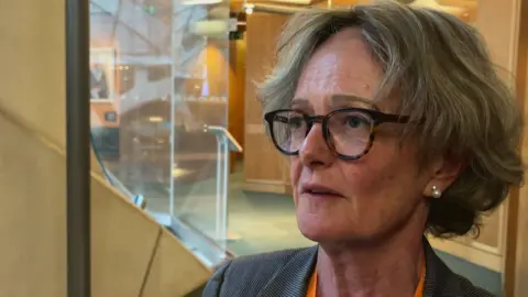 A middle aged woman with glasses, light coloured hair and a pearl earring speaks to a reporter while standing in a modern office building foyer.