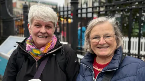 Two women - Kim McMahon and Julie Dean - stand outside the front gates of Belfast City Hall and smile at the camera. They are both older women, with Kim having short, white hair, and Julie having slightly longer grey hair. Both wearing are wearing puffed coats.
