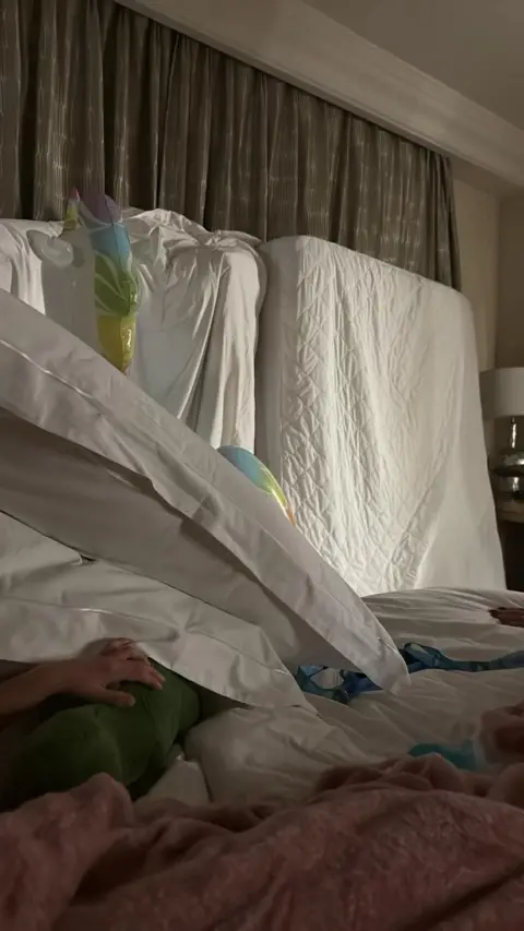 Hollie Hill Multiple mattresses up against a window in a hotel room. The patterned curtains are closed. Mattresses also line the floor.