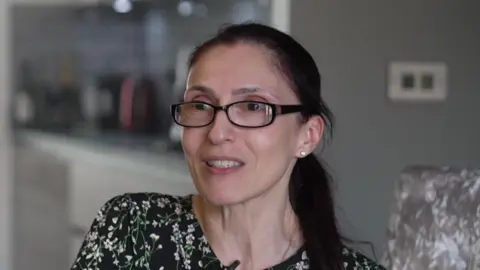 A woman with long black hair wearing black rectangular glasses smiles looking away from the camera. She is wearing a black top with floral pattern. 