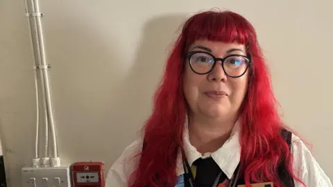 Joolz Bailey standing against a white wall. She has long pinky red hair and is wearing glasses, a white shirt and over it a black sleeveless top. She is carrying a clipboard and starting to smile. 