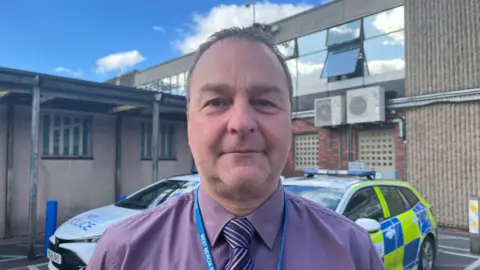 Det Insp Davies looking into the camera; he is wearing a purple collared shirt with a purple, pink, and black striped tie. He has got a blue lanyard around his neck with West Mercia Police on it in white writing. Behind him are two marked police cars, and beyond that is Shrewsbury Police Station - a square, brutalist grey brick building with large windows. The sky is blue with a few clouds. 