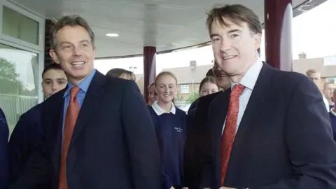 Tony Blair, in navy blue suit and red tie, stands alongside Peter Mandelson, in similar attire, out on a campaign event in 2001. There are some school children behind them as they walk into a school in Mandelson's Hartlepool constituency, which he left the same year. 