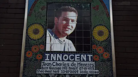Getty Images Memorial mosaic of Jean Charles de Menezes, featuring his portrait framed by colorful flowers and the word "INNOCENT" in bold letters beneath. The text below includes his birth details and states he was "sadly missed".