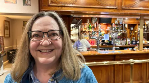 Jennifer Barton with long blonde hair, wearing a blue sweater and sitting in front of the club bar, which is made of wood and is behind a gold-coloured railing. Bottles, optics and a cash register are visible at the bar, and a person in a grey top is preparing a drink.