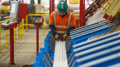 Bloomberg via Getty Images A manufacturing worker holds steel rods in a production line next to buttons and machinery while wearing an orange reflective jacket and green helmet, at the Marcegaglia Steel SpA bar finishing unit in Sheffield in March 2025.