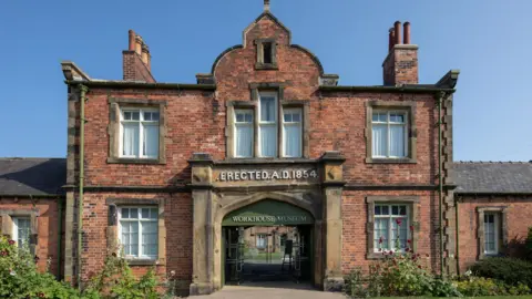 A brick building with an archway in the middle.  The signs on the building say "Erected A.D. 1854" and "Workhouse Museum".