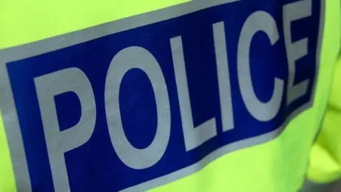 A close-up of the back of a police officer's high visibility jacket showing the word "police" in silver letters on a dark blue background.