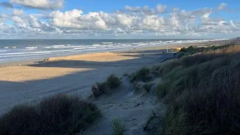 A photograph of the beach at De Panne in Belgium, which shows small sand dunes in the foreground, then a sandy stretch of beach and the Channel coast beyond that.