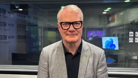 A man with white hair and dark-framed glasses, wearing a grey suit and black shirt, stands in a radio studio. Beyond him a large internal glass window and computer screens can be seen.