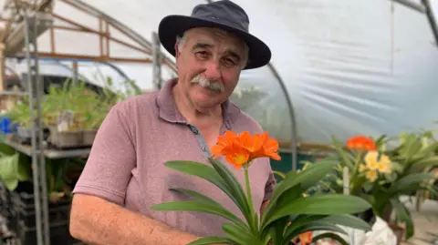 A man in a hat and dark pink T-shirt holds an orange flower with several flower heads. In the background, shelves with plants can be seen.
