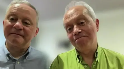 A screenshot of a Zoom interview with Fr Martin O'Hagan and Fr Eugene O'Hagan. Fr Martin is on the left, with short grey hair and wearing a blue, buttoned-up shirt. On the right is Fr Eugene with grey hair, wearing a green, button-up shirt.