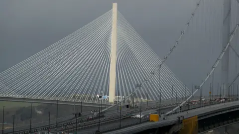 The Queensferry Crossing suspension bridge stands behind the Forth Road Bridge with a grey sky in the background. 