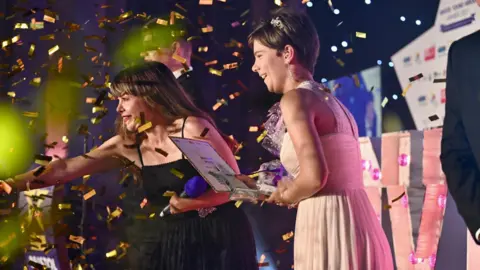 Dougie Allward/Made For Impact Host Briony Williams is at left with dark hair, wearing a black dress and pointing into the audience while Overall Hero Chloe is on stage in a blush evening gown smiling with her prizes - she has short dark hair and is also looking in the direction of the audience