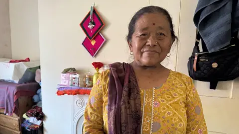 A Gurkha widow is wearing a bright yellow dress with a red shawl draped over her right shoulder. She also has a gold stud in her left nostril. Staring straight into the camera she's sitting in her bedroom and you can see the vague outlines of a few glass jars over her shoulder as well. A beautiful deep red shawl thrown over her right shoulder Indumati Thapa is standing in her small bedroom. Wearing a yellow dress you can see the foot stool she sits on when not in her bed behind her. 